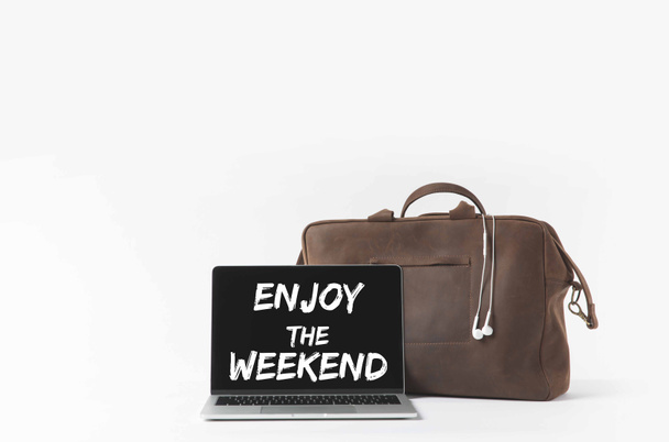laptop met geniet van het weekend belettering op scherm en stijlvolle tas met koptelefoon, geïsoleerd op wit - Foto, afbeelding