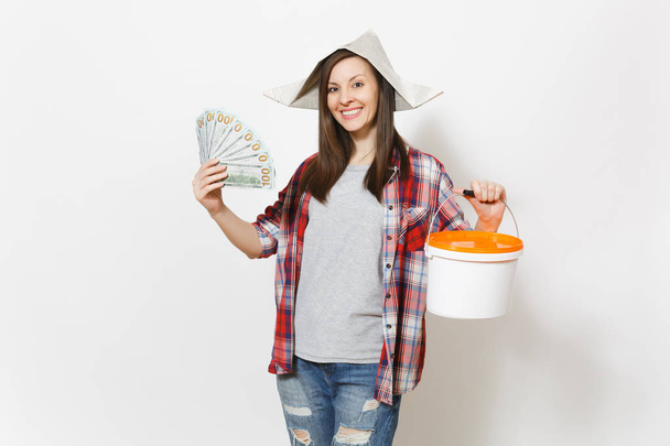 新聞ドルの持株のバンドルを帽子の女性を笑顔現金お金と空のペイントはバケツ白い背景に分離されたコピー スペース付き。リノベーション マンションの楽器。修理ホーム コンセプト. - 写真・画像