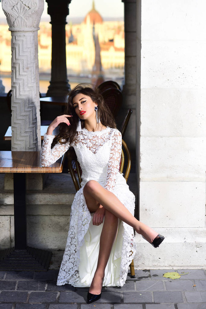 Fashion model with glamour look and makeup. Sexy woman sit alone in street cafe. Woman in white wedding dress outdoor. Girl with long brunette hair. Bride with makeup face - Fotoğraf, Görsel