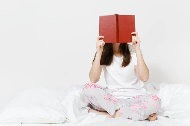 Young happy woman sitting in bed with white sheet, pillow, blanket on white background. Beauty female spending time in room, reading book, hiding behind book. Relax, good mood concept. Place for text. - Zdjęcie, obraz