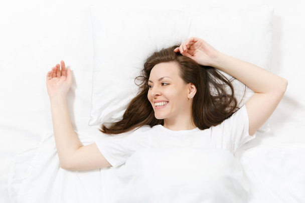 Draufsicht auf den Kopf der glücklichen brünetten jungen Frau, die mit weißem Laken, Kissen, Decke im Bett liegt. lächelnde hübsche Frau, die Zeit im Zimmer verbringt. Ruhe, Entspannung, gute Laune. Kopierfläche für Werbung - Foto, Bild