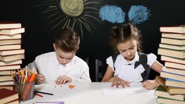School concept. Cute children enjoy ssuccess sitting at a desk with heaps of books and a blackboard with drawings behind them. High five. Goal reach. 4k - Footage, Video