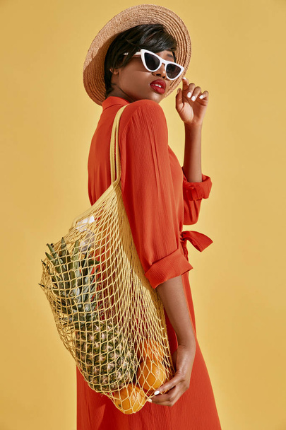 Studio portrait of beautiful young black woman with short brown hair, red lips, dressed in orange red dress, white sunglasses, hat, with fruits in net bag. Yellow background - Foto, Bild
