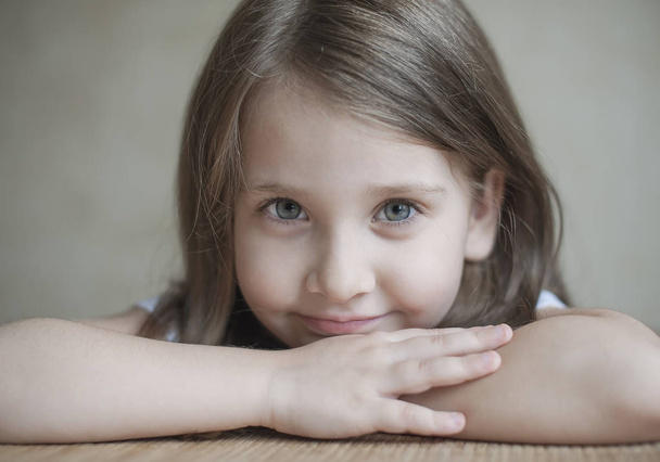 A little smiling girl is sitting at the table, looking at the camera. Emotional portrait. Curiosity, drive, energy, enthusiasm, inquiry, interest, joy, laugh, loving, mischief, pleasure, tenderness - Fotografie, Obrázek