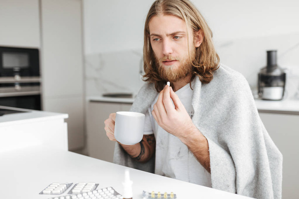 Retrato de homem doente sentado com caneca e pílulas nas mãos na cozinha em casa isolado
 - Foto, Imagem
