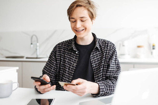 Портрет щасливого чоловіка в навушниках, що сидять з банківською картою та мобільним телефоном в руці на кухні вдома ізольовані
 - Фото, зображення