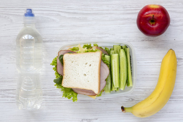 gesunde Schulessenbox mit frischem Bio-Gemüse-Sandwich, Obst und einer Flasche Wasser auf weißem Holzgrund, flach gelegt. von oben. - Foto, Bild