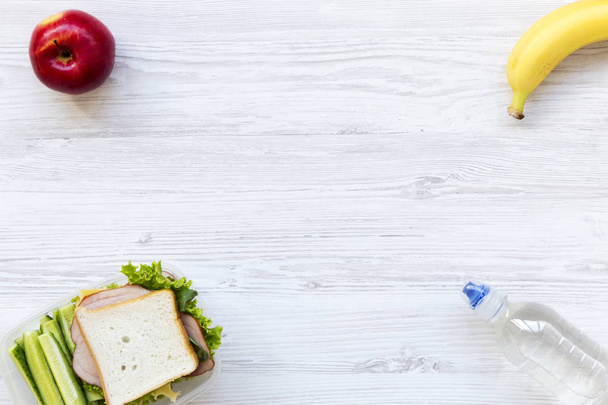 gesunde Schulessenbox mit Sandwich, Früchten und Wasserflasche auf weißem Holzgrund, flach gelegt. von oben. Kopierraum. - Foto, Bild