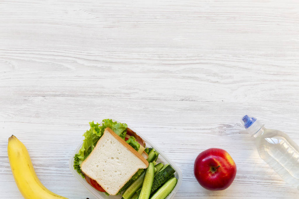 Школьная обеденная коробка с бутербродами, фруктами и бутылкой воды на белом деревянном фоне, плоский уголок. Сверху. Копирование пространства
. - Фото, изображение