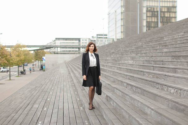 European businesswoman standing on stairs with bag and high buildings in background. - Fotografie, Obrázek