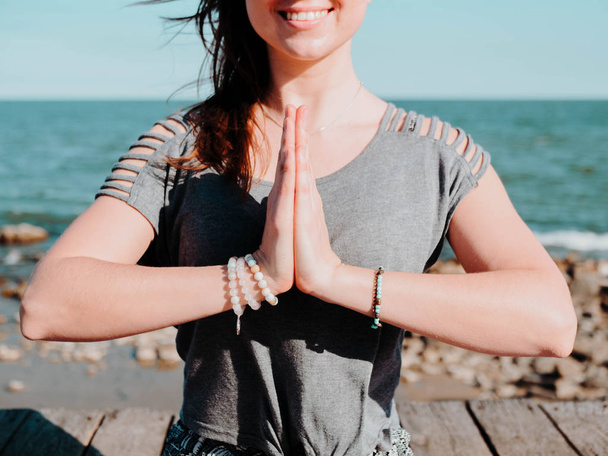 Young beautiful sporty woman doing yoga mudra on sea wooden beach near water. Girl practicing exercises. Hands in namaste. Gratitude sign. Health concept. - Fotoğraf, Görsel