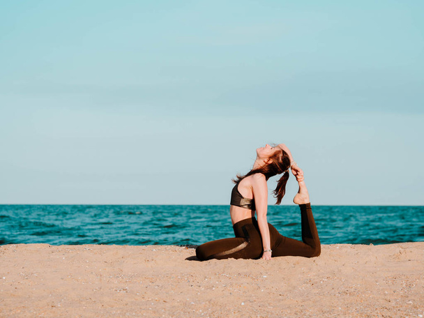 Young beautiful sporty woman in green clothing doing yoga asana on sea sandy beach near water. Girl practicing exercises. Health concept. Copy space. - Foto, afbeelding