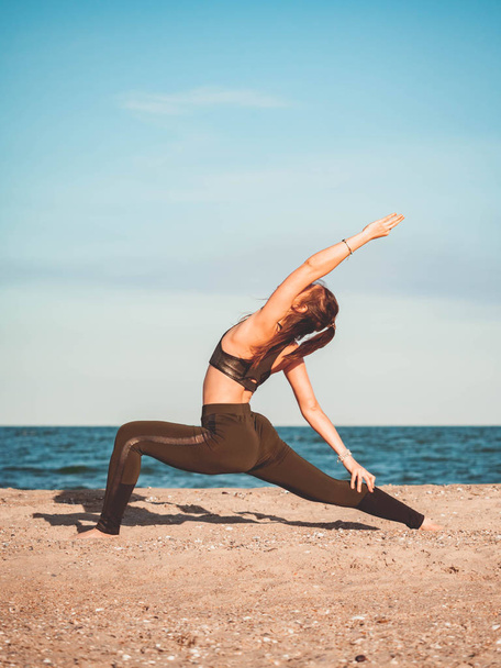 Young beautiful sporty woman in green clothing doing yoga asana on sea sandy beach near water. Girl practicing exercises. Health concept. Copy space. - Photo, image