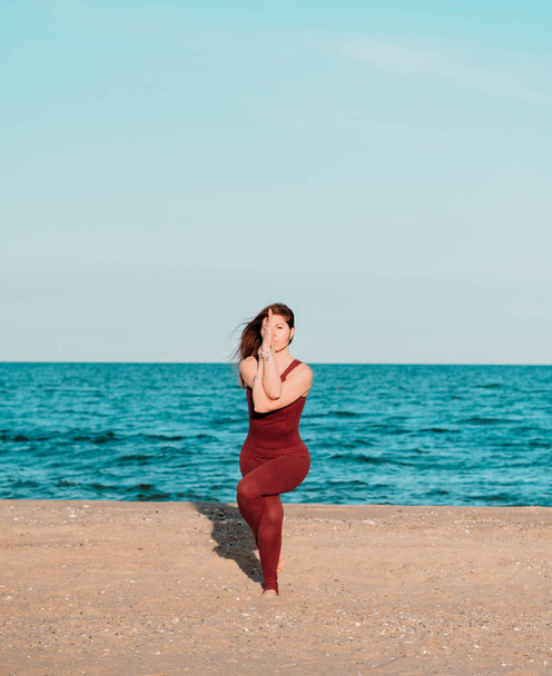 Young beautiful sporty woman in red costume doing yoga asana on sea beach near water. Girl practicing exercises. Health concept. - Foto, immagini