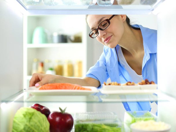 Portrait of female standing near open fridge full of healthy food, vegetables and fruits - Photo, Image