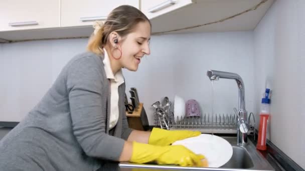 4 k πλάνα του happy χαμογελαστή γυναίκα ακούγοντας μουσική με ακουστικά ενώ τα πλένοντας πιάτα στην κουζίνα - Πλάνα, βίντεο