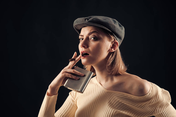 Blonde lady looks like suspicious detective. Detective concept. Girl thinking about investigation, holds flask and smoking cigar, copy space. Woman on mysterious face wears kepi, dark background - Фото, изображение