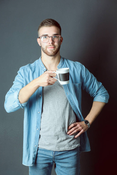 Portrait of a handsome young man standing and holding a cup of coffee in his hands - Zdjęcie, obraz