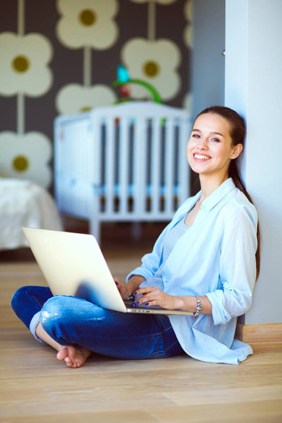 Young woman sitting on the floor near childrens cot with laptop. Young mom - Foto, Imagen