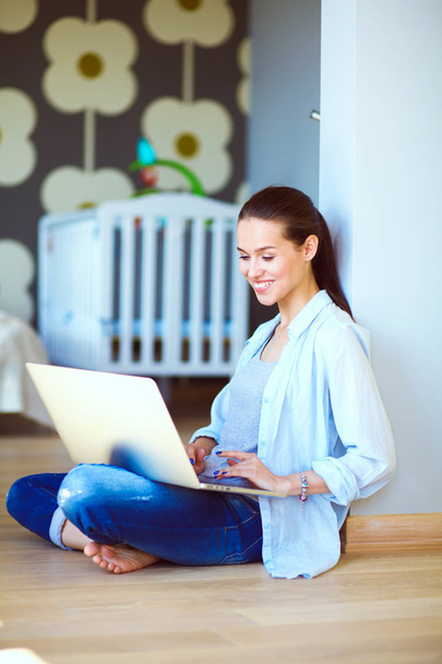 Young woman sitting on the floor near childrens cot with laptop. Young mom - Photo, Image