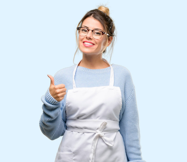 Власниця магазину молодої жінки, одягнена в фартух, посміхається широко показуючи великі пальці вгору жестом до камери, виражаючи як і схвалення
 - Фото, зображення