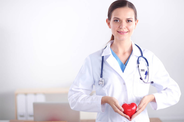 Beautiful young smiling female doctor sitting at the desk and holding heart. - Фото, изображение