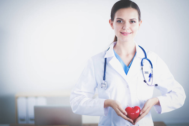 Beautiful young smiling female doctor sitting at the desk and holding heart. - Foto, afbeelding