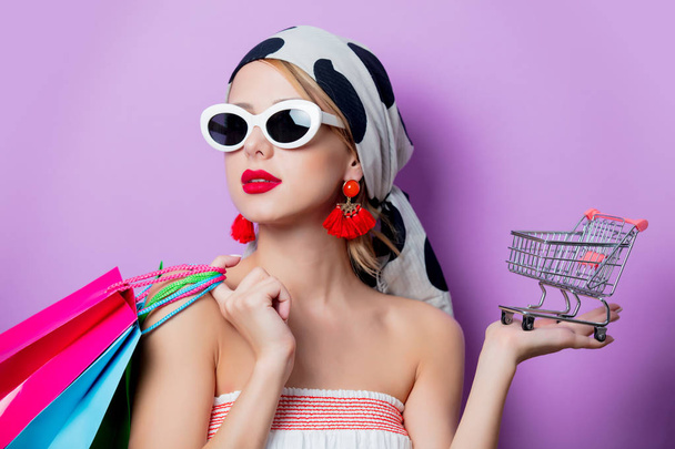 Retrato de mujer de estilo hermoso en gafas de sol con carrito pequeño y bolsas de compras sobre fondo púrpura
 - Foto, Imagen