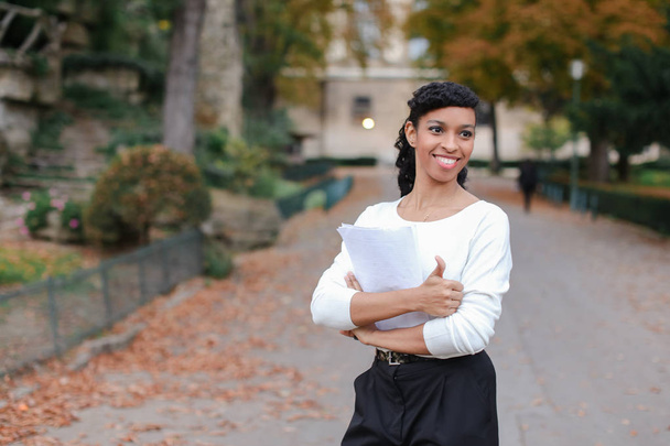 Αφρο αμερικανικό όμορφου φοιτητή περπάτημα με έγγραφα στο πάρκο φθινόπωρο, δείχνει τους αντίχειρες επάνω. - Φωτογραφία, εικόνα