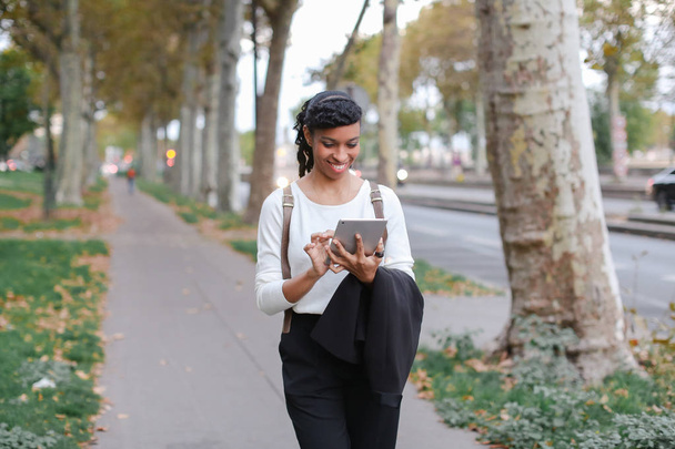 Μαύρο όμορφη φοιτήτριας χρησιμοποιώντας tablet και το περπάτημα στο δρόμο με τα δέντρα. - Φωτογραφία, εικόνα