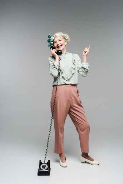 smiling senior woman talking by vintage phone and pointing up on grey - Fotó, kép