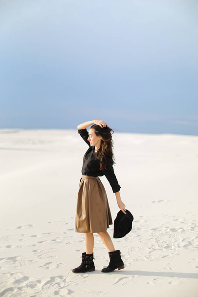 Young pretty female photo model standing on snow and wearing skirt and black blouse. keeping hat. - Photo, Image