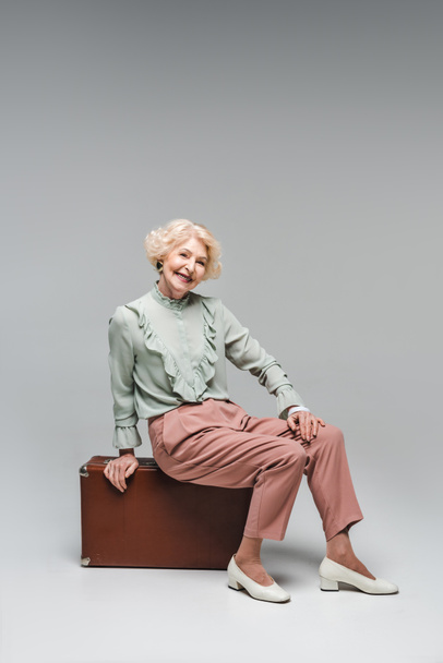 beautiful senior woman sitting on vintage suitcase on grey - Foto, Imagem