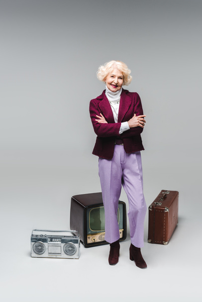 beautiful senior woman with crossed arms standing in front of vintage tv, boombox and suitcase on grey - Foto, Imagem