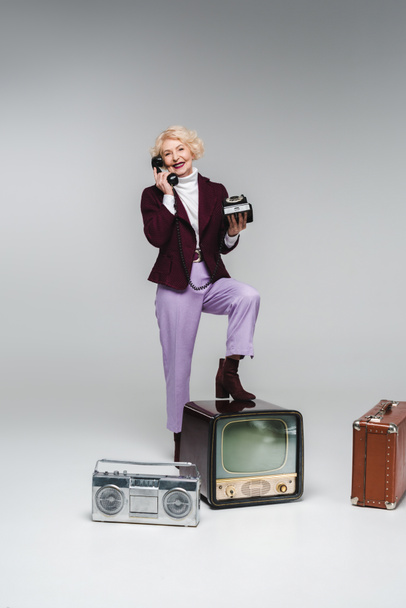 hermosa mujer mayor hablando por teléfono mientras está de pie en la televisión vintage en gris
 - Foto, Imagen