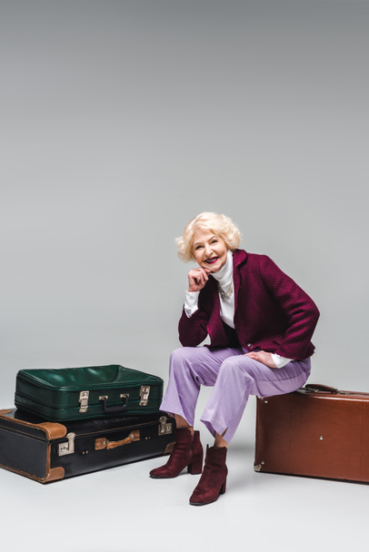 happy senior woman sitting on vintage suitcase on grey - Foto, immagini