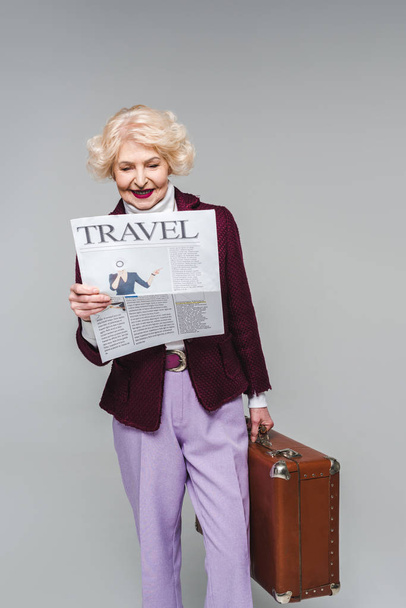 όμορφο ανώτερων γυναίκα κρατάτε βαλίτσα και διαβάζοντας εφημερίδα ταξιδιού που απομονώνονται σε γκρι - Φωτογραφία, εικόνα