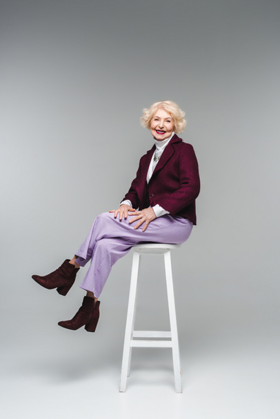 attractive senior woman in stylish clothes sitting on chair on grey - Zdjęcie, obraz