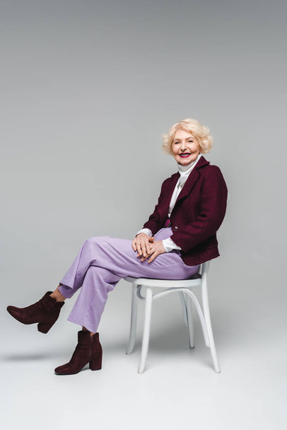 mujer mayor sonriente sentada en la silla y mirando a la cámara en gris
 - Foto, imagen