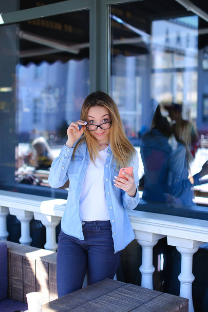 Chica bonita tocando gafas y manteniendo el teléfono inteligente en la cafetería
. - Foto, Imagen