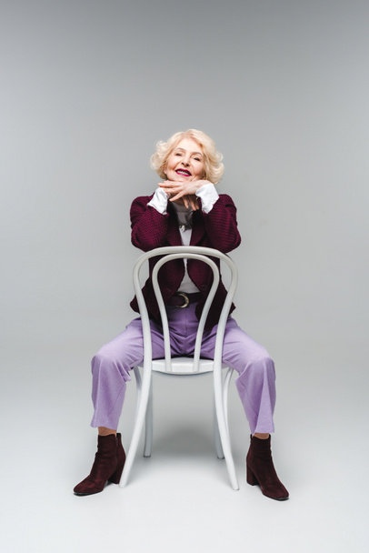 happy senior woman sitting on chair and looking at camera on grey - Φωτογραφία, εικόνα