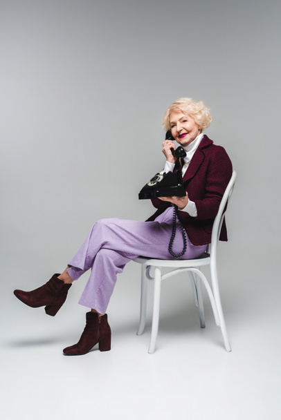 beautiful senior woman talking by vintage phone while sitting on chair on grey - Photo, Image