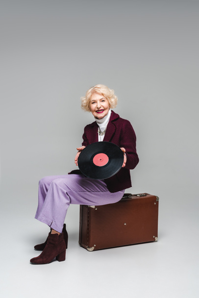 happy stylish senior woman with vinyl disc sitting on vintage suitcase on gray background  - Foto, Bild