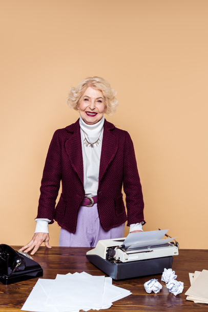 sonriente mujer mayor con estilo de pie en la mesa con máquina de escribir y teléfono giratorio
 - Foto, Imagen