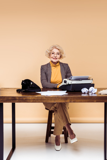 smiling stylish senior woman using typewriter at table with papers and rotary phone - Foto, Imagen