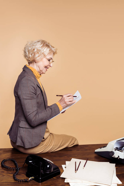 side view of stylish senior woman writing on paper and sitting on table with typewriter and rotary phone - Foto, imagen