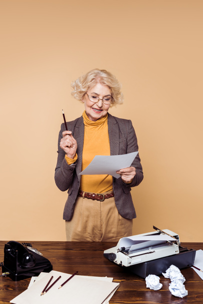 Stilvolle Seniorin mit Brille blickt mit Schreibmaschine und Drehtelefon auf Papier am Tisch - Foto, Bild