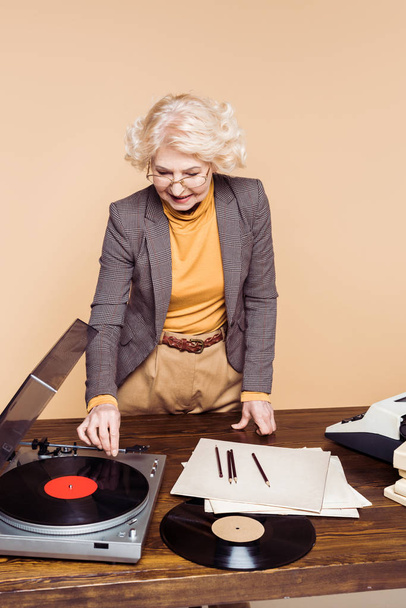 mujer elegante senior que enciende el reproductor de discos de vinilo en la mesa con máquina de escribir y disco de vinilo
 - Foto, Imagen