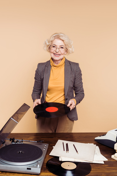 レコード プレーヤーのテーブルの近くのビニール ディスクを保持しているスタイリッシュなシニア女性  - 写真・画像