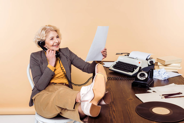 mujer mayor sonriente con las piernas en la mesa hablando por teléfono rotatorio y mirando pape
 - Foto, Imagen